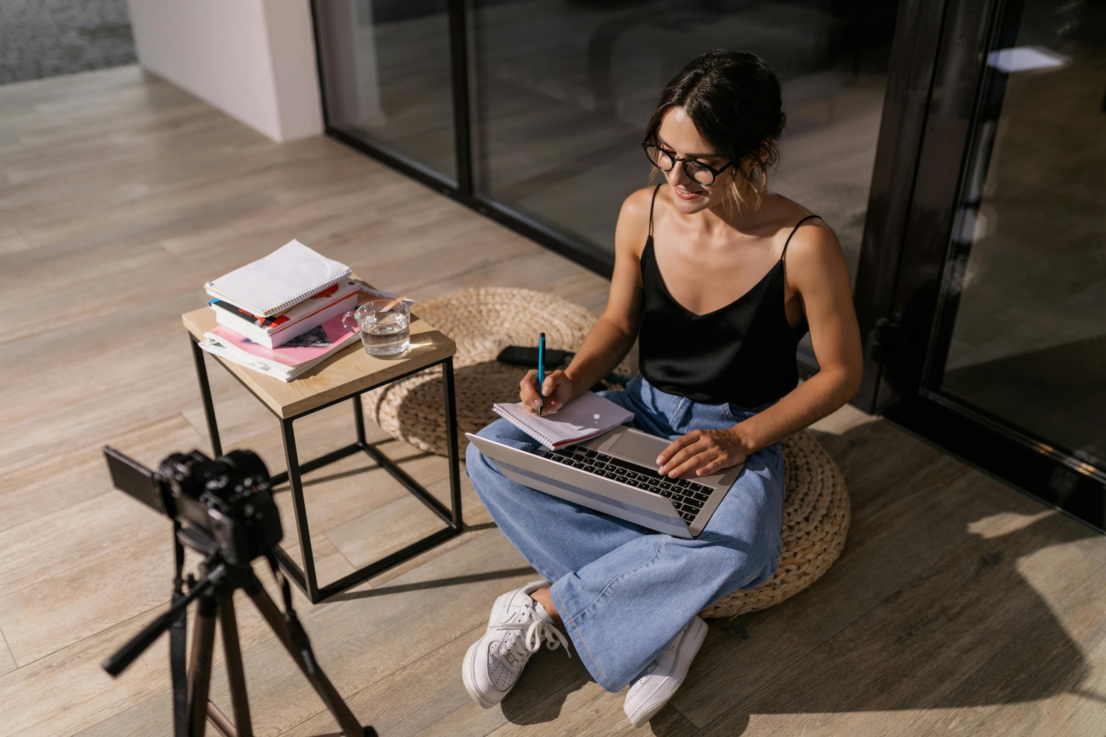 A woman sits on a cushion, working on a laptop and taking notes for an online course. A camera is set up on a tripod in front of her, and a small table beside her holds books and a glass of water.