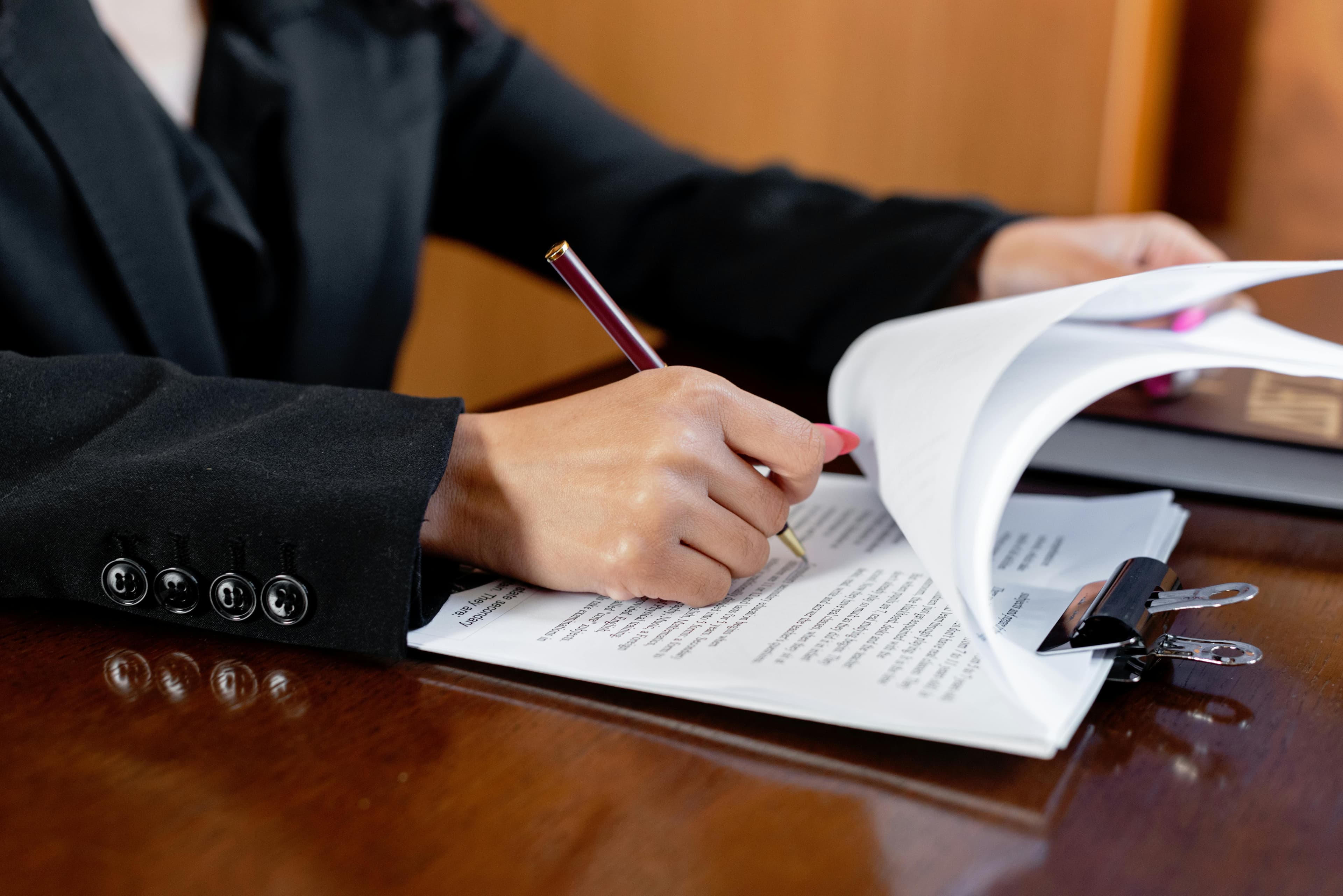 A person in a black suit writes with a pen on a stack of documents clipped together on a wooden table. The content focuses on legal content and con-compete clauses for businesses. 