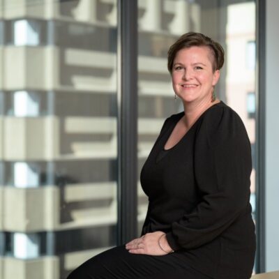 Professional woman with short hair smiles, sitting by a window in an office setting.